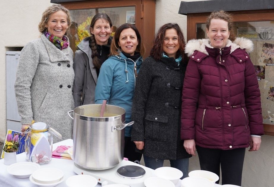5 Frauen stehen mit winterlicher Kleidung im Freien. Im Vordergrund steht ein Tisch mit Tellern und ein großer Suppentopf