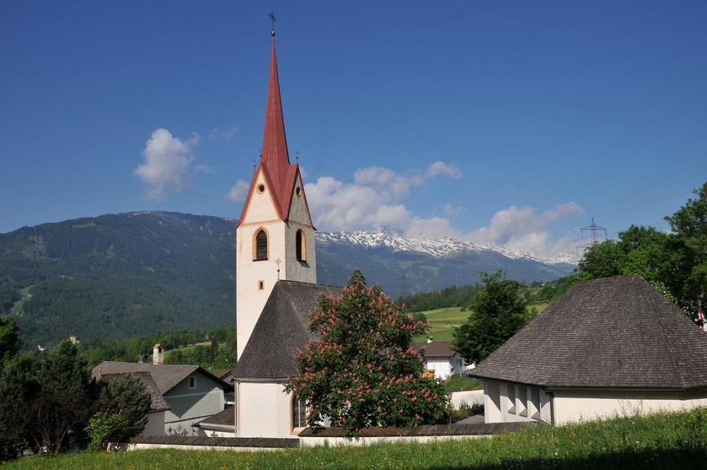 Pfarrkirche umgeben von Häusern und grünen Wiesen vor blauem Himmel. Bergrücken im Hintergrund.