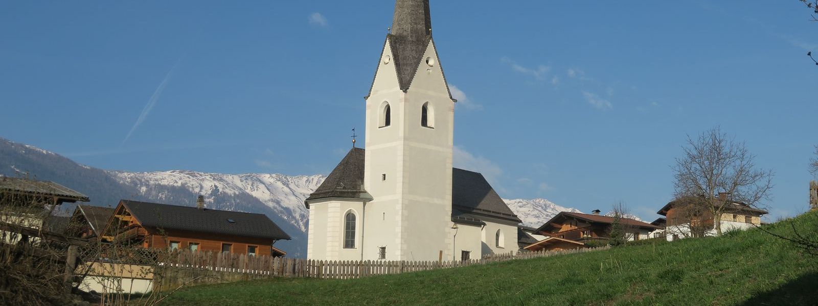 Kirche vor einer grünen Wiese vor blauem Himmel. Dahinter schneebedeckte Berge
