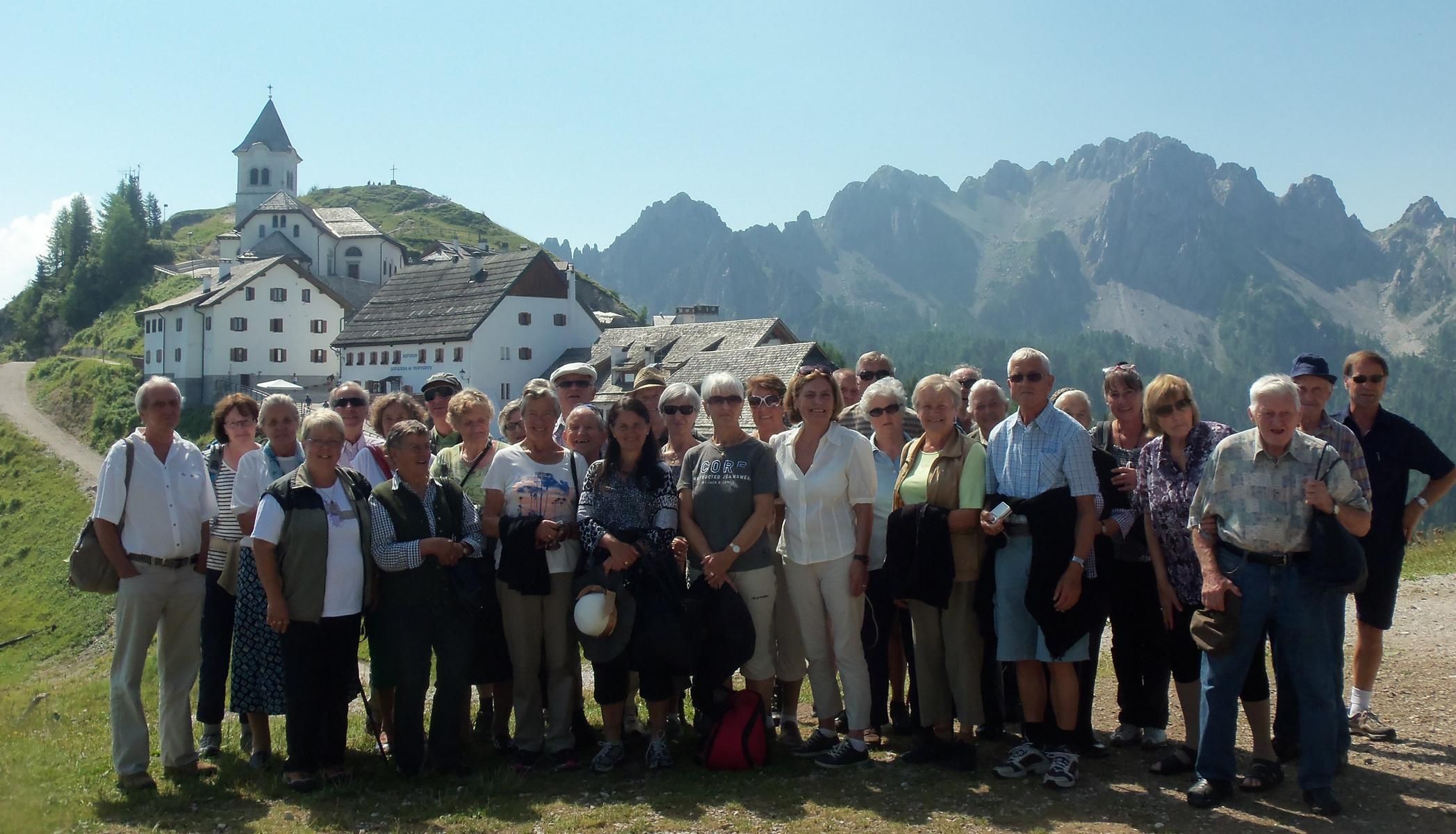 Eine Gruppe von Senioren stehen bei blauem Himmel auf einer Wiese. Im Hintergrund sieht man eine Kirche.