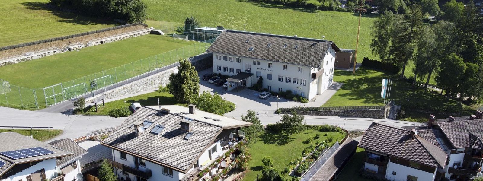 Luftaufnahme vom Gemeindehaus. Links daneben der Sportplatz. Im Vordergrund einige Häuser der Gemeinde