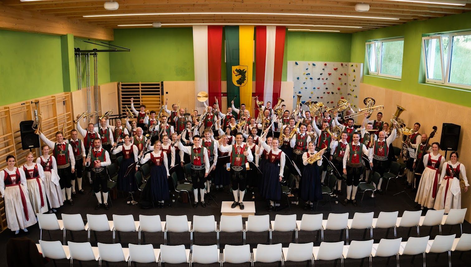 Die Mitglieder der Musikkappelle sitzen und stehen in Tracht gekleidet in 4 Reihen hintereinander.
