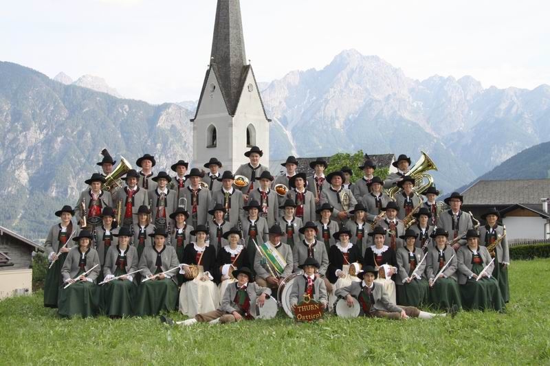 Bild der Musikkapelle auf einer Wiese. Dahinter der Kirchturm vor einer Bergkette