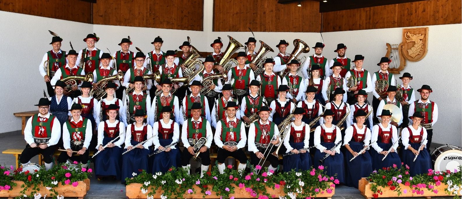 Die Mitglieder der Musikkappelle sitzen und stehen in Tracht gekleidet in 4 Reihen hintereinander.